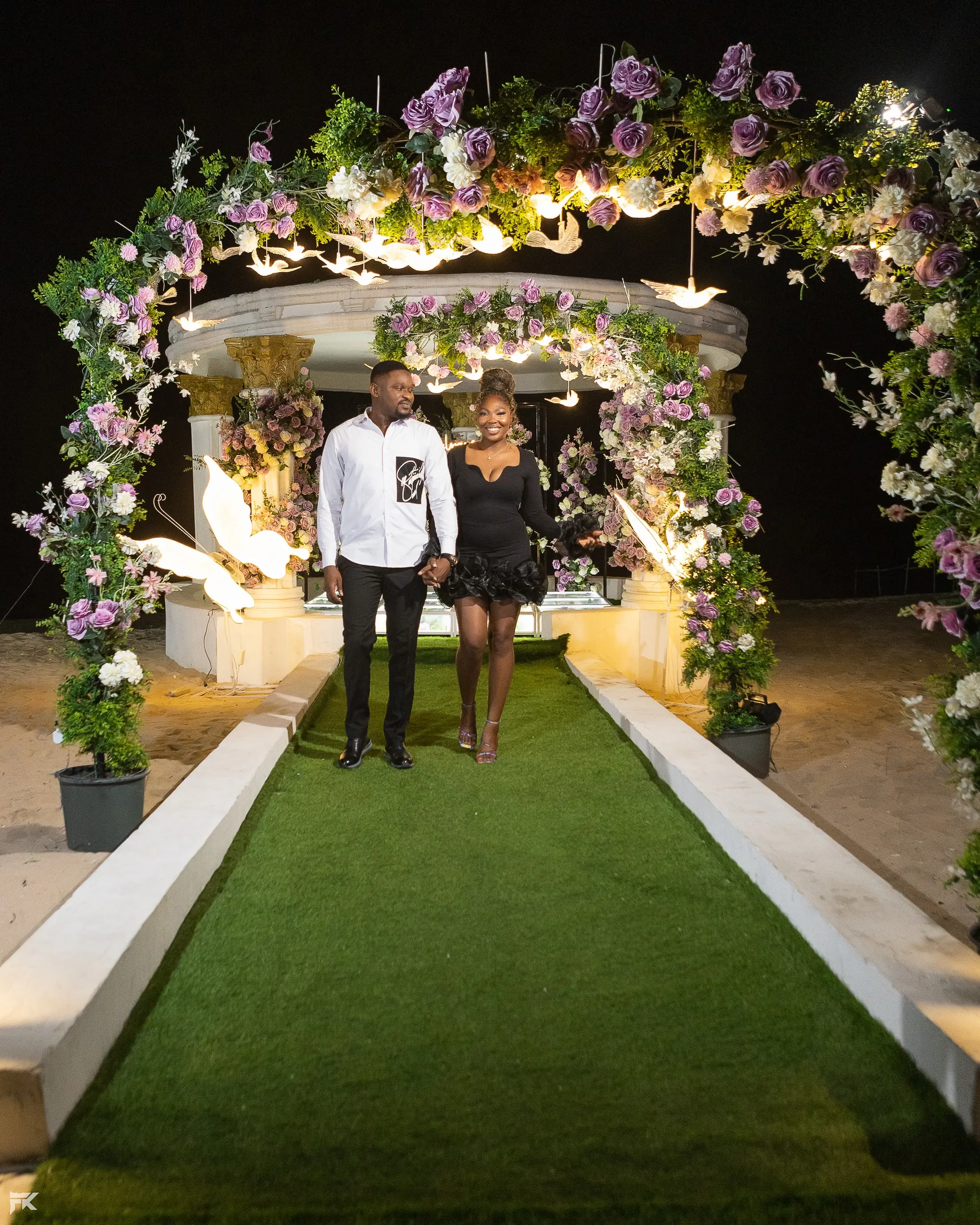 A couple standing under a lit flower arch at the end of a green runway at night