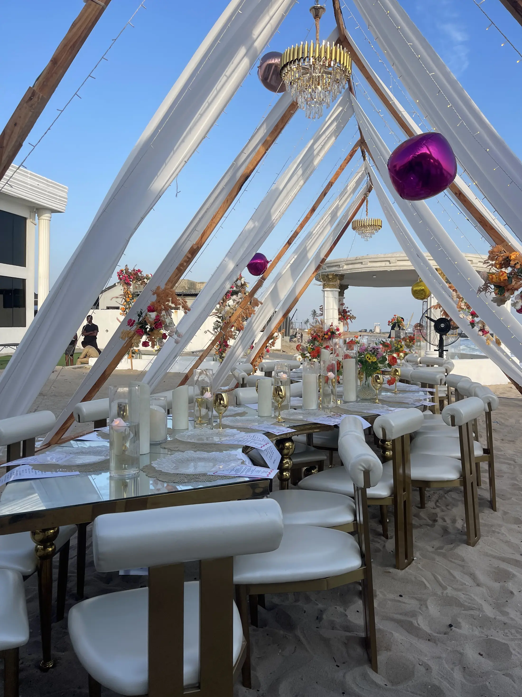 A long dinner table on the sand under white draped A-frames in daylight
