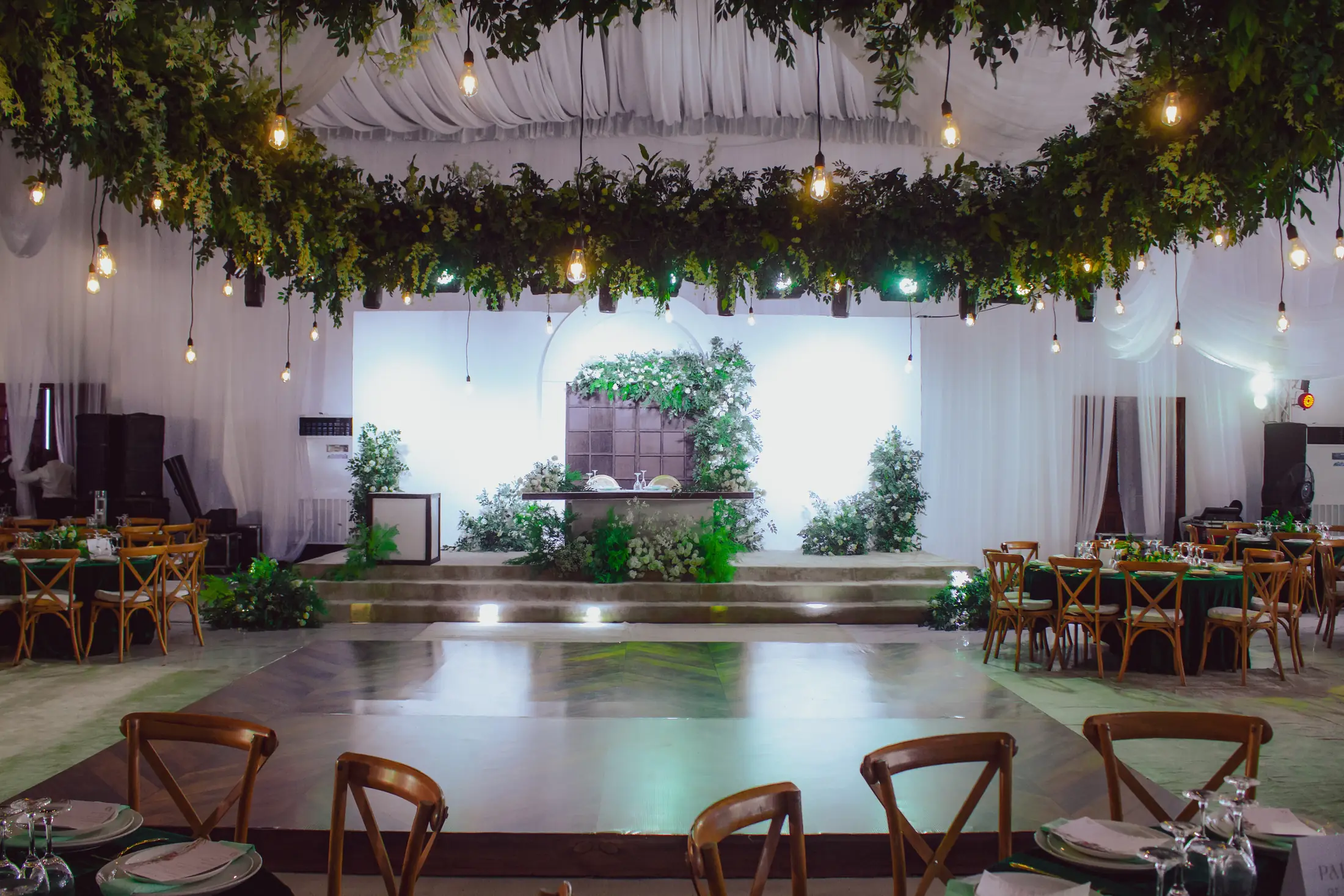 The hall dressed for a reception under hanging greenery and bulb lights, a polished dance floor before the stage