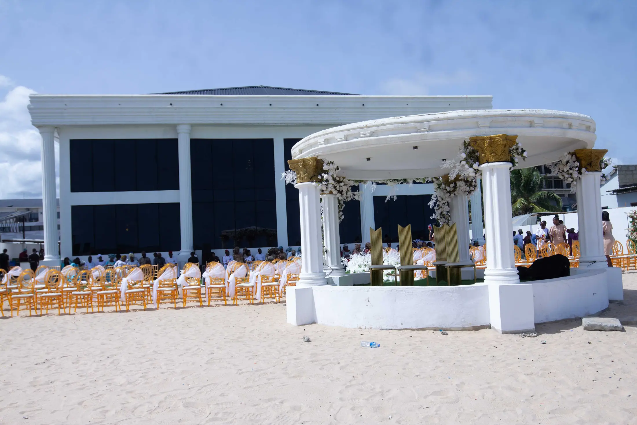 The white colonnaded hall behind the gazebo on the sand, guests seated for a ceremony