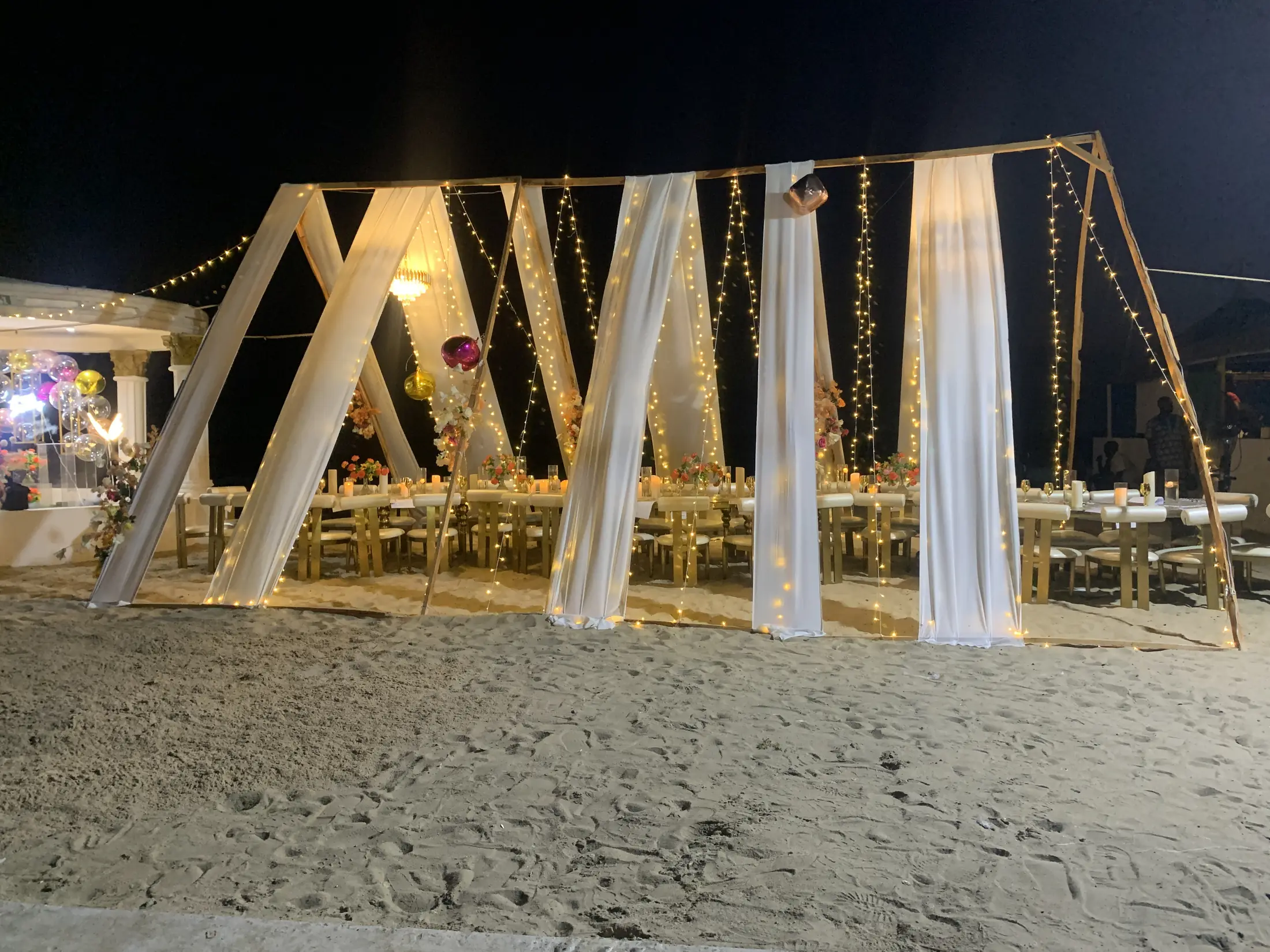 White draped A-frames lit with string lights over long tables on the sand at night