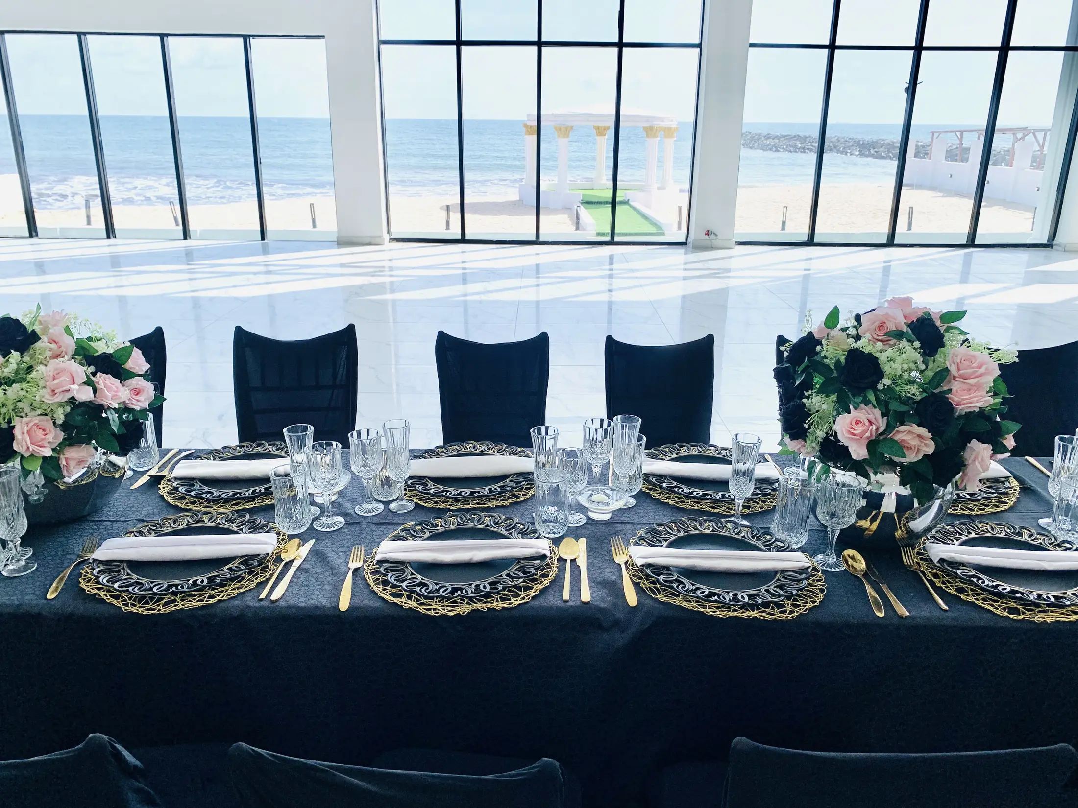 A long dinner table dressed in black and gold against the hall's glass wall, the beach and sea beyond