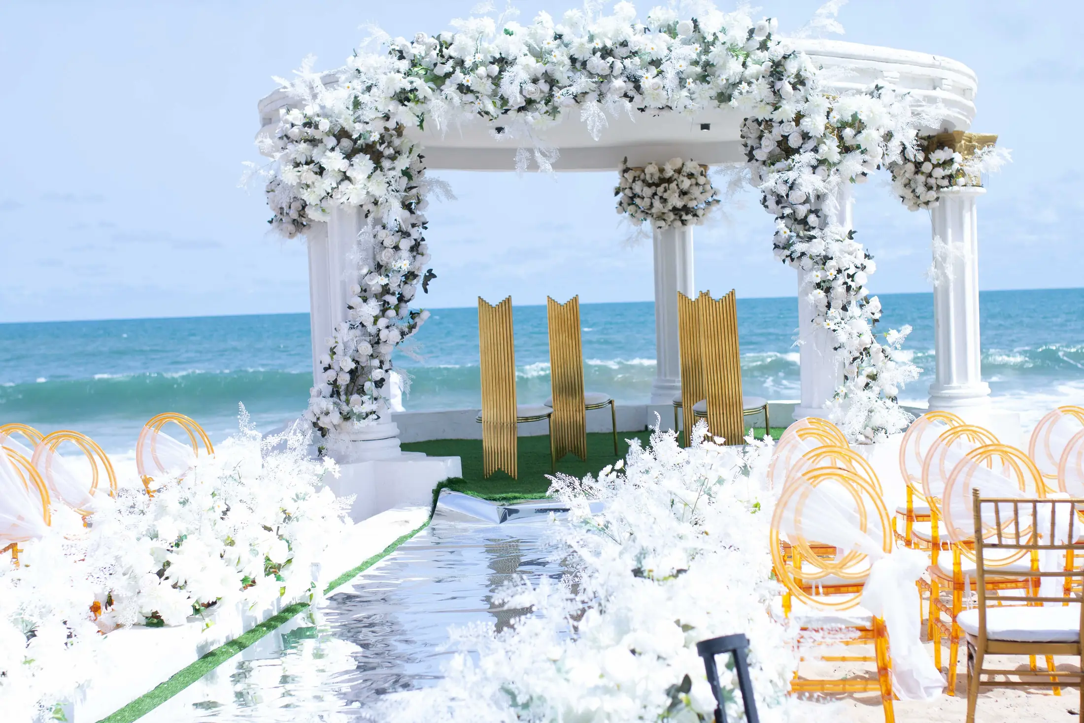 The gazebo dressed in white flowers, gold chairs facing it, the sea behind