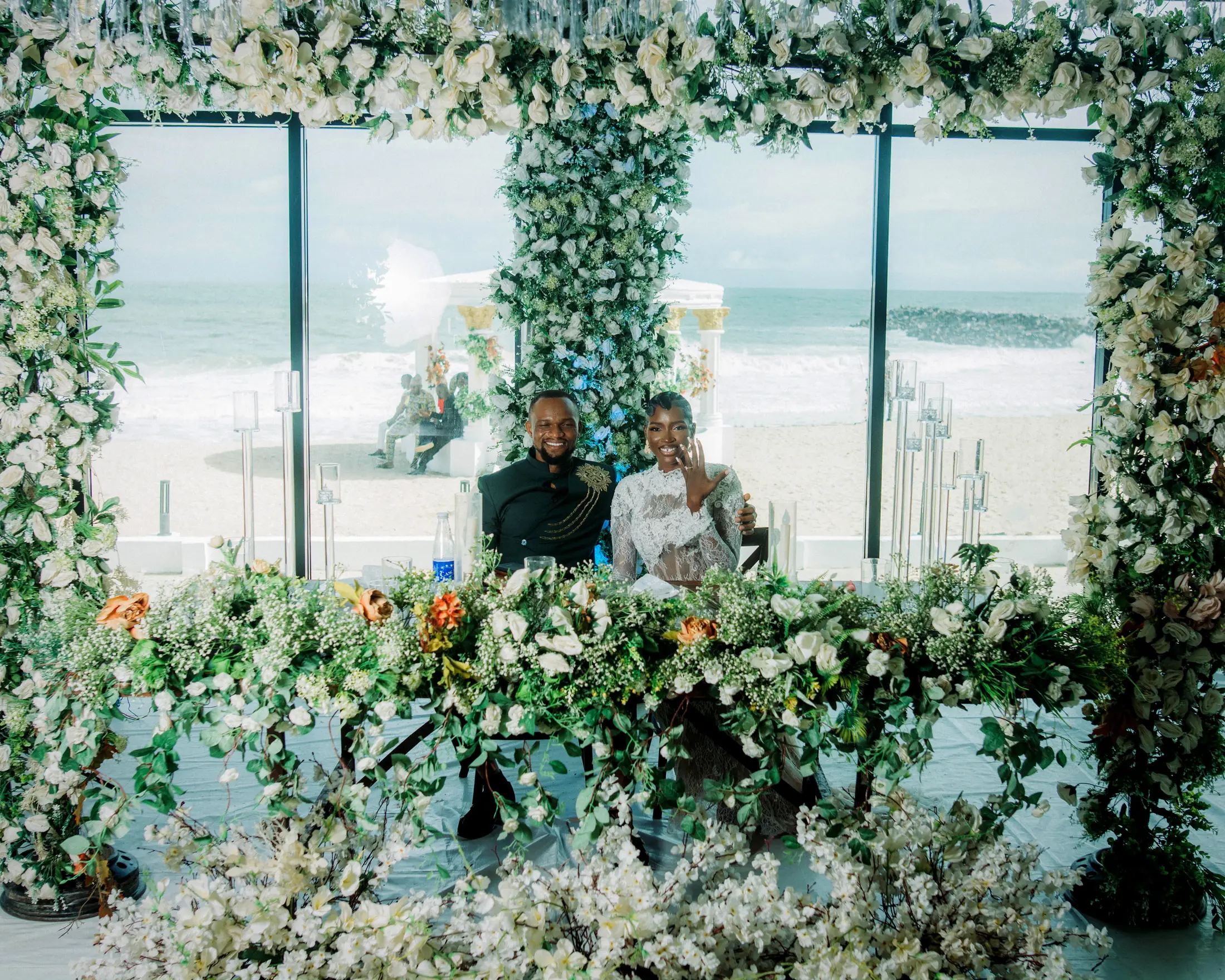 A bride and groom smiling at their flower-covered table, the sea through the glass behind them