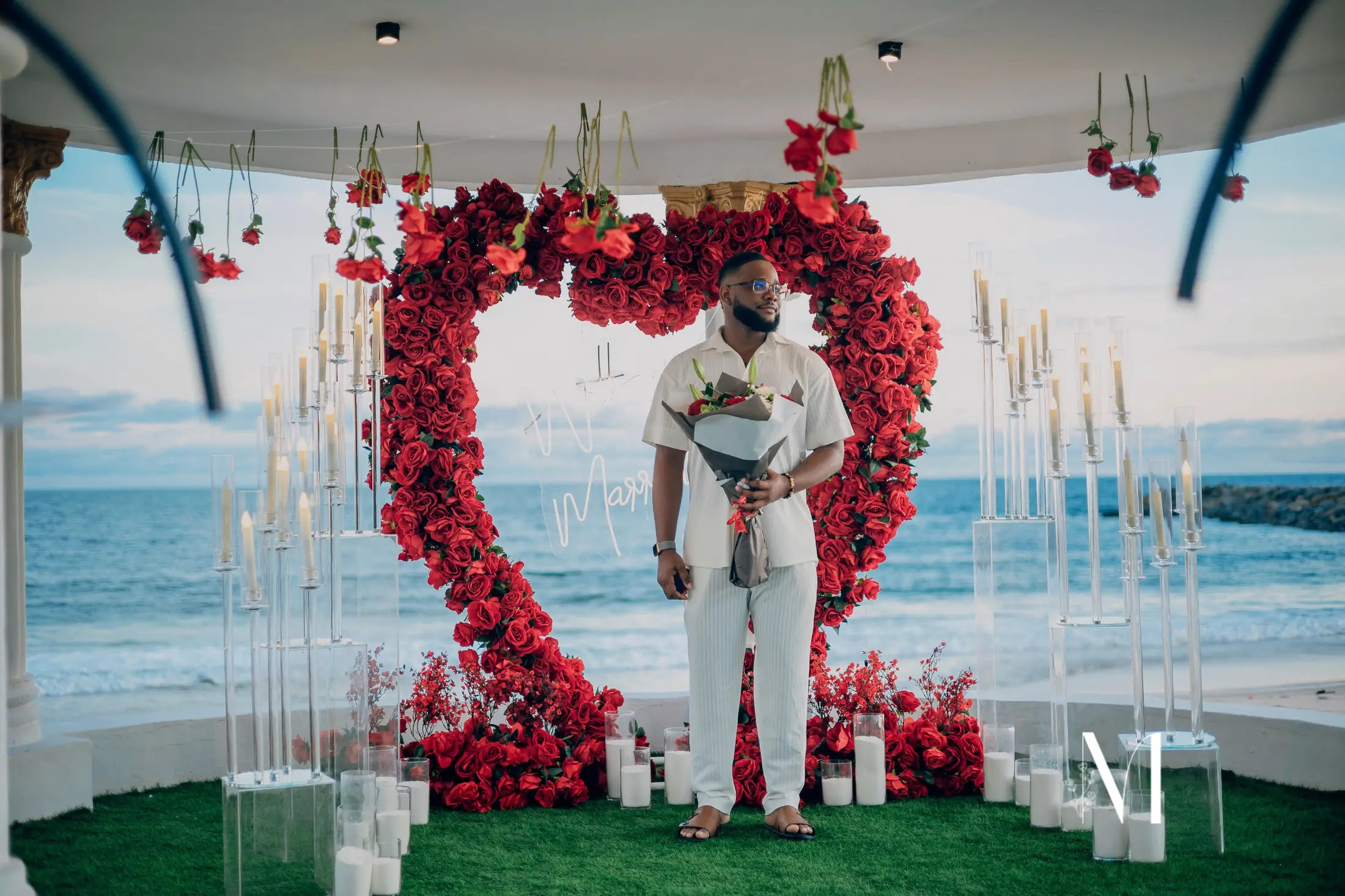A man waiting inside a heart of red roses by the sea, holding flowers
