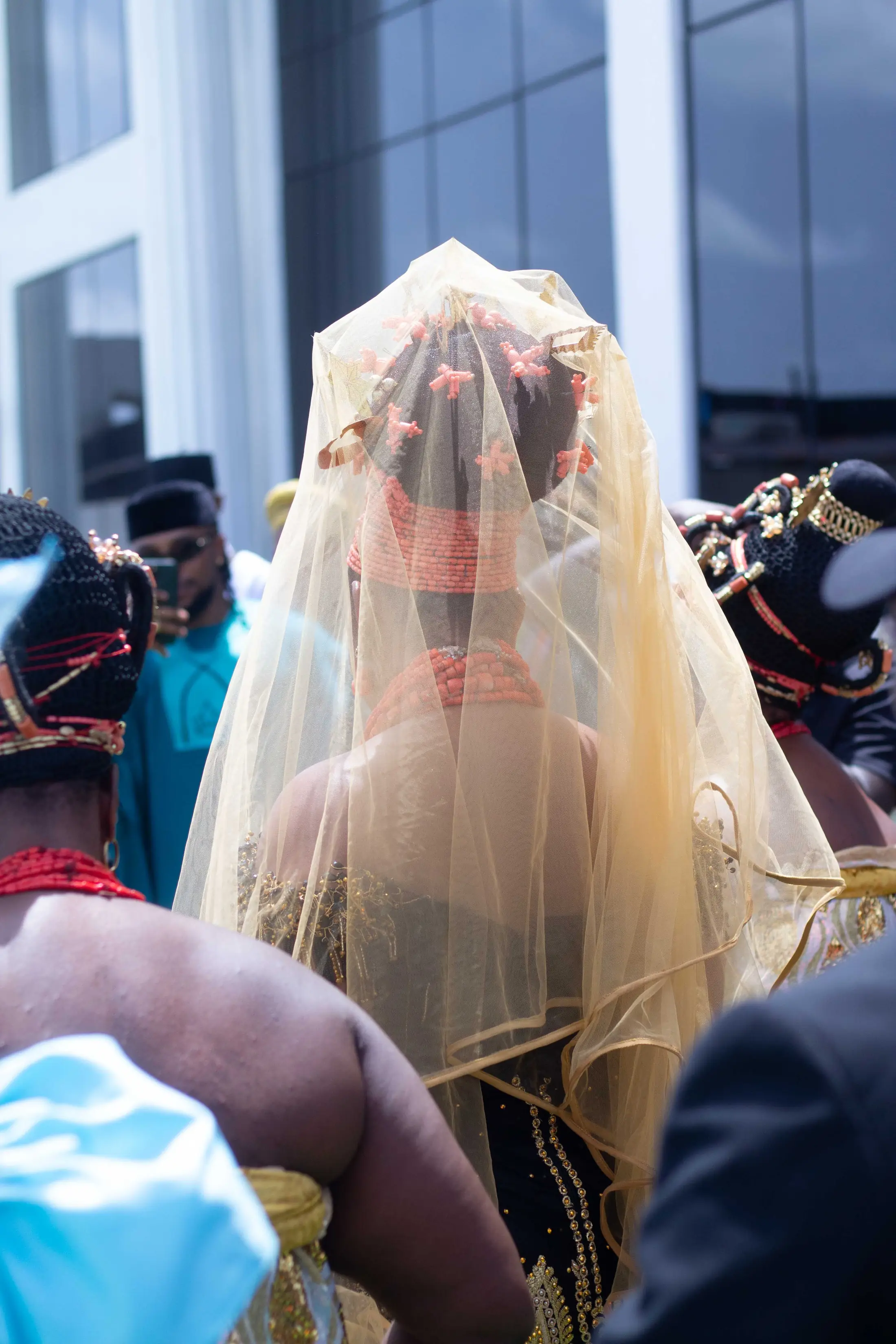 A bride in coral beads looking down, her veil lit by the sun