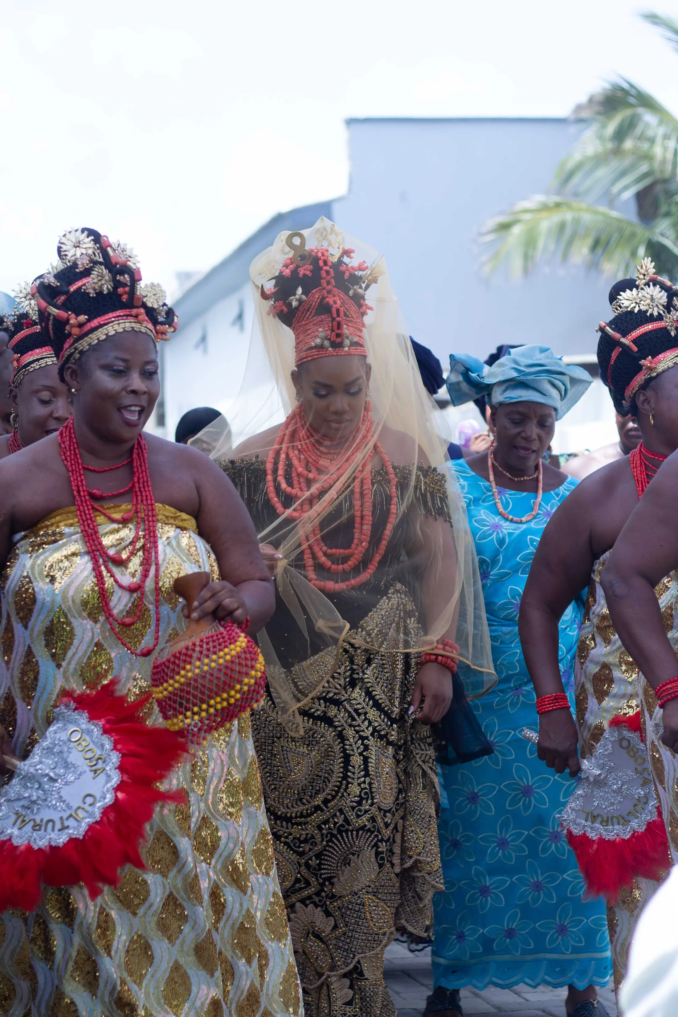 Women in coral beads and gold wrappers dancing in with red feather fans